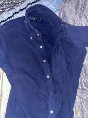 Blå skjorta från Ralph Lauren - Säljer en klassisk blå skjorta från Ralph Lauren med button-down krage och vita knappar. Skjortan är i bomull och har en tidlös design som funkar till många olika stilar. Perfekt för dig som gillar en clean och enkel look. Den var för liten för mig så säljer det billigt, saknar även en knapp men annars är allt bra inga hål eller sånt. 