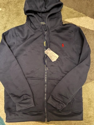 Marinblå Polo Ralph Lauren hoodie - Marinblå hoodie från Polo Ralph Lauren med dragkedja, huva och snörning. Klassisk roterad logga på bröstet och känguruficka framtill. Tillverkad i ett mjukt bomullsmaterial, perfekt för en clean och stilren look.