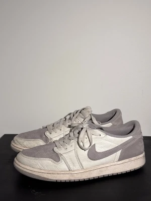 Nike Jordan 1 Low i grått och vitt - Nike Jordan 1 Low sneakers i en clean färgkombo av ljusgrått och vitt. Skorna har klassisk snörning, rund tå och platt sula. Ovandelen är i skinn och mocka med meshfoder för extra komfort. Swoosh-loggan på sidan och Nike Air-märke på plösen.