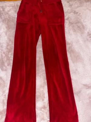 Juicy Couture röda mjukisbyxor - Snygga röda mjukisbyxor från Juicy Couture i mjuk sammet med bred resår och snörning i midjan. Byxorna har raka ben, stora fickor med metallknappar och broderad JC-logga på fickan. Perfekt för chill eller streetwear.