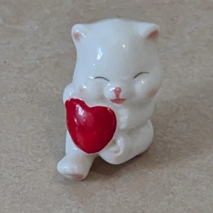 Söt vit kattfigur med rött hjärta - Supergullig liten vit katt som håller ett rött hjärta. Perfekt som dekoration eller present till någon du gillar. Katten har rosa detaljer på nos och öron och ett glatt ansiktsuttryck. Hjärtmotivet gör den extra charmig och unik.
