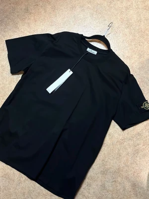 Svart t-shirt från Stone Island - Svart t-shirt från Stone Island med klassisk rund hals och korta ärmar. T-shirten har den ikoniska Stone Island-loggan på vänster ärm och är gjord i mjuk bomull. Enkel och stilren design som funkar till allt. Perfekt för dig som gillar streetwear.