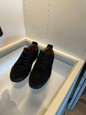 Christian Louboutin svarta spikes sneakers - Säljer ett par svarta sneakers från Christian Louboutin i mocka med ikoniska röda sulor. Skorna har svarta nitar över tån, svarta snören och en clean, låg siluett. Perfekta sop sneakers för dig som vill sticka ut.