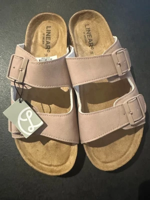 Ljusa rosa sandaler från Linear - Snygga sandaler i ljusrosa från Linear of Sweden med två breda remmar och justerbara spännen. Ovandelen är i mockaliknande material och sulan är vit med mönstrad undersida för extra grepp. Perfekta för varma dagar och chill vibes.