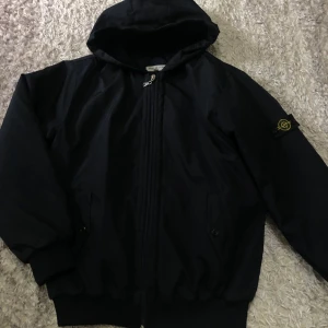 Svart vindjacka med huva från Stone Island - Säljer en svart vindjacka från Stone Island med huva och dragkedja framtill. Jackan har ribbade muddar, två fickor med knappar och den klassiska Stone Island-loggan på ärmen. Perfekt för dig som gillar stilrena och sportiga plagg.