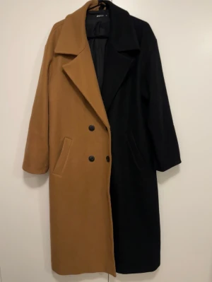 Tvåfärgad lång kappa från GINATRICOT - Säljer en riktigt snygg lång kappa från Ginatricot i tvåfärgad design – ena halvan är svart och andra halvan är brun. Kappan har dubbelknäppning, klassisk krage och raka ärmar. Perfekt statement-plagg för höst och vinter.
