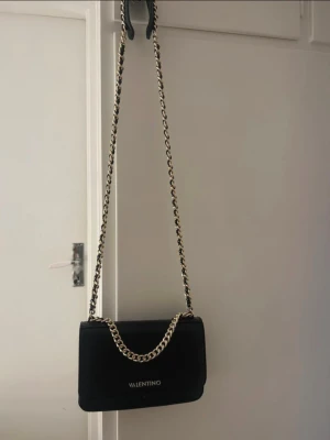 Svart axelväska från Valentino - Snygg svart axelväska från Valentino med guldfärgad kedja och logga framtill. Väskan är i slät skinnimitation och har ett stilrent, fyrkantigt format. Invändigt finns dragkedjefack och smarta fack för småsaker. Nypris 1200kr