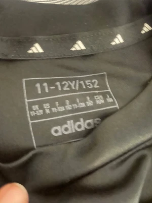 Svart Adidas t-shirt med vita ränder - Snygg svart Adidas t-shirt med klassiska vita ränder längs axlarna och Adidas-logga på bröstet. Tillverkad i mjukt syntetmaterial som andas, perfekt för sport eller chill. Passar dig som gillar stilren och sportig look.