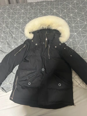 Svart Moose Knuckles dunjacka med päls - Svart dunjacka från Moose Knuckles med fluffig vit päls runt huvan, flera dragkedjor och snygga detaljer. Jackan har broderad insida med hockeymotiv och praktiska fickor. Perfekt för kalla dagar och har en cool streetstil.