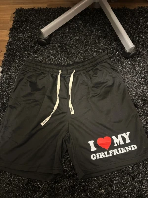 Svarta shorts med I ❤️ MY GIRLFRIEND-tryck - Svarta shorts med vit snörning i midjan och stort tryck på ena benet där det står 'I ❤️ MY GIRLFRIEND' i vitt och rött. Shortsen har elastisk midja och är tillverkade i ett mjukt, syntetiskt material. Perfekta för chill dagar eller som statement-plagg. De har även fickor. De är helt oanvända