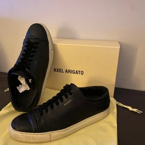 Svarta sneakers från Axel Arigato - Snygga svarta sneakers från Axel Arigato med klassisk rund tå och vit platt sula. Skorna är tillverkade i slätt skinn och har svarta snören. Minimalistisk design med diskret guldfärgad logga på sidan. Perfekta för dig som gillar stilrena och trendiga sneakers.