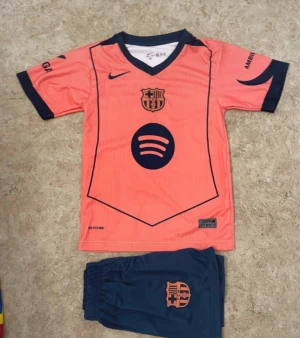 Barça Lamine Yamal fotbollströja set - Säljer ett Barcelona fotbollströja och shorts-set med Lamine Yamal #10. Tröjan är korallrosa med mörkblå detaljer, klubbmärke och sponsortryck. Shorts är mörkblå med korallrosa klubbmärke. Perfekt för unga fans som vill sticka ut på planen.