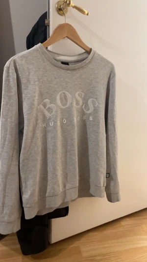 Grå sweatshirt från Hugo Boss - Snygg grå sweatshirt från Hugo Boss med stort BOSS-tryck i vitt på bröstet. Tröjan har rund hals, långa ärmar och ribbade muddar vid ärmslut och nederkant. Perfekt för en chill och stilren look.
