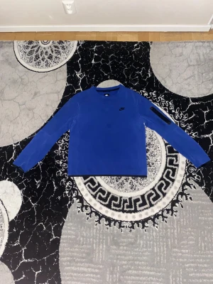 Blå långärmad Nike tröja S - Säljer en blå långärmad tröja från Nike i storlek S. Tröjan har en stilren design med rund hals och ett diskret Nike-logga på bröstet. Materialet känns mjukt och är troligtvis polyester, perfekt för träning eller chill. Ärmarna har en svart detalj på ena sidan.