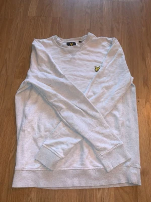 Grå sweatshirt från Lyle & Scott - Vit sweatshirt från Lyle & Scott med klassisk rund hals och gul broderad logga på bröstet. Tröjan har ribbade muddar vid ärmslut och nederkant samt långärmad passform. Perfekt för dig som gillar stilrena och enkla plagg.