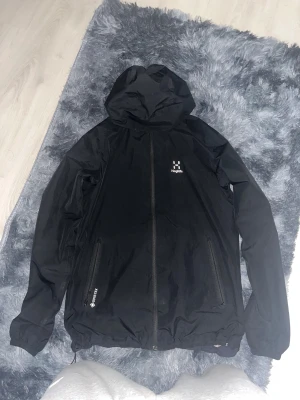 Svart Haglöfs vindjacka Gore-Tex - Säljer en svart vindjacka från Haglöfs med huva och hel dragkedja framtill. Jackan är tillverkad i slitstark Gore-Tex och har två dragkedjeförsedda fickor. Perfekt för dig som vill ha en clean och funktionell look.