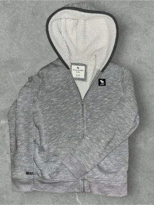 Abercrombie & Fitch zip-hoodie – grå - Abercrombie & Fitch zip-hoodie – grå  Mjuk och bekväm zip-hoodie från Abercrombie & Fitch i gråmelerad färg. Insida i vit teddy/fleece som gör den extra varm och skön. Hel dragkedja framtill, huva med mjukt foder samt ribbade muddar vid ärmslut och nederkant. Diskret logga på bröstet och broderad text på ärmen.  Storlek: 13–14 år Passar även: XS/S (jag är 178 cm och den sitter nästan bra) Skick: Mycket bra Passform: Normal / något mindre S/XS