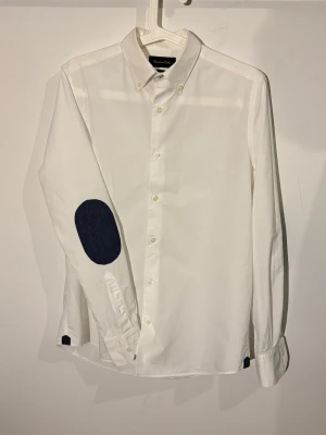 Massimo Dutti armbågslappar - Snygg vit skjorta från Massimo Dutti med mörkblå armbågslappar och klassisk krage. Skjortan har knappar framtill och långa ärmar med detaljer i mörkblått vid manschetterna. Perfekt för dig som gillar stilrena plagg med en twist.