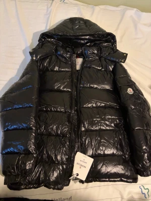 Svart pufferjacka från Moncler M - Säljer en svart, glansig pufferjacka från Moncler i storlek M. Jackan har huva, dragkedja framtill och klassisk Moncler-logga på ärmen. Perfekt för kalla dagar och riktigt snygg streetstil. Materialet är syntet och dun, vilket gör den både lätt och varm.