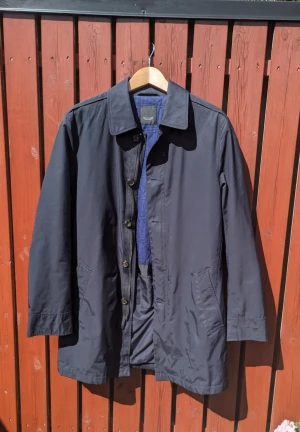 SAND Copenhagen, trenchcoat, mörkblå, bomull, nylon, läder - Lätt fodrad, klassisk mörkblå trenchcoat från Sand Copenhagen i en väderrobust bomull/nylon-blandning. Perfekt i lager på lager till vår/höst. Läcker läderdetalj i nacken. Ordinarie pris c:a 3800 kr. I mycket gott skick och använd vid ett fåtal tillfällen.  Bröstmått (armhåla till armhåla): 110 cm. Längd (krage till fåll): 86 cm.  Vid frågor kontakta mig gärna via chatt eller email.