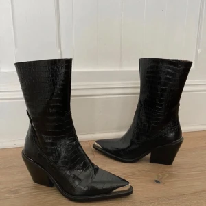 Svarta cowboy boots med spetsig tå - Säljer ett par svarta cowboy boots från Zara med croco-mönstrad yta i skinn med metall-detalj. Storlek 38 men passar även en 37. I superfint skick! 