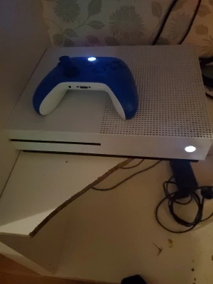 Xbox One S - Säljer en Xbox One S i vitt med tillhörande blå/vit handkontroll. Konsolen är i gott skick och fungerar som den ska, inga synliga defekter eller större slitage. Perfekt för gaming och streaming. Strömkabel och HDMI-portar synliga på bilderna. Handkontrollen lyser och är testad.