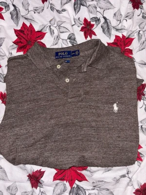 Mörkgrå piké från Polo Ralph Lauren - Snygg mörkgrå pikétröja från Polo Ralph Lauren med klassisk krage och två knappar framtill. Tröjan har det ikoniska broderade logotypen i vitt på bröstet och är tillverkad i mjuk bomull. Perfekt för en clean och stilren look.