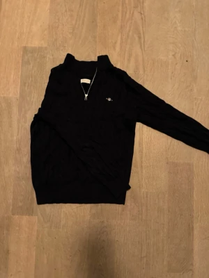 Mörk blå stickad tröja med half zip från Gant - Svart stickad tröja från Gant med halv dragkedja och diskret logga på bröstet. Tröjan har lång ärm, ribbade muddar och klassisk kabelstickad struktur. Perfekt för dig som gillar stilren och tidlös design.