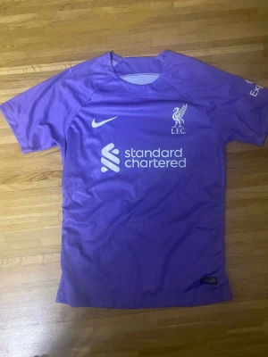 Lila Liverpool Nike matchtröja - Säljer en lila Liverpool FC matchtröja från Nike med vita loggor och tryck. Tröjan har kort ärm, rund hals och är tillverkad i ett lätt, syntetiskt material som andas. Perfekt för dig som älskar fotboll och vill sticka ut på planen eller läktaren.