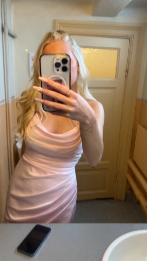 Ljusrosa långklänning - Säljer en elegant ljusrosa långklänning med snygg drapering och cowlneck. Klänningen är ärmlös och har en figurnära passform som framhäver siluetten. Materialet är glansigt och känns lyxigt, perfekt för student eller bal. Passar dig som vill sticka ut med en feminin och stilren look.