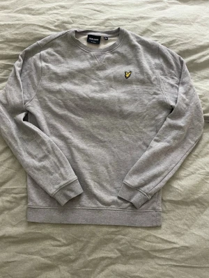 Grå sweatshirt från Lyle & Scott - Säljer en stilren grå sweatshirt från Lyle & Scott med klassisk gul broderad logga på bröstet. Tröjan har rund halsringning, långa ärmar och ribbade muddar. Perfekt för dig som gillar enkel och tidlös stil.