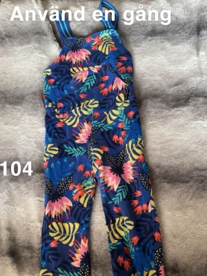 Blommig byxdress i blått och rosa - Färgglad byxdress med blommigt mönster i blått, rosa, gult och grönt. Byxdressen har breda axelband och en stor ficka framtill. Perfekt för dig som gillar lekfulla och unika plagg med mycket färg.🌸 Använda en gång. Storlek 104