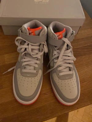 Nike Air Force 1 Mid grå/orange 42 - Nike Air Force 1 Mid '07 i grått med detaljer i ljusgrått och stark orange på plös, häl och yttersula. Klassisk snörning, kardborreband vid ankeln och perforerad tåbox. Snygg kontrast mellan den neutrala ovandelen och den färgstarka sulan. Perfekt för dig som vill sticka ut.