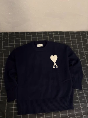 Mörkblå stickad Ami Paris tröja - Säljer en mörkblå stickad tröja från Ami Paris med ett vitt hjärta och bokstaven A broderat på bröstet. Tröjan har rund hals, ribbade muddar och är långärmad. Perfekt för dig som gillar stilrena detaljer och vill ha något som sticker ut lite extra. Bara testad på, i nyskick. Inga defekter, hör av dig om du har några frågor.