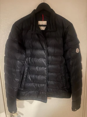  Moncler jacka  - Säljer en svart pufferjacka från Moncler med klassisk quiltad design och Moncler-logga på ärmen. Jackan har dragkedja framtill, ståkrage och praktiska fickor med knappstängning. Perfekt för kalla dagar och riktigt snygg till streetwear.