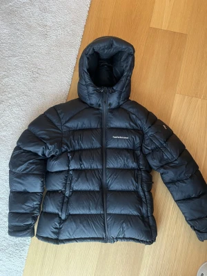 Mörkblå Peak Performance Frostdown jacka - Säljer en mörkblå/svart pufferjacka från Peak Performance med huva och dragkedja framtill. Jackan har en quiltad design och är perfekt för kalla dagar. Klassisk logga på bröstet och praktiska fickor på sidorna. Passar dig som vill ha en varm och stilren vinterjacka.