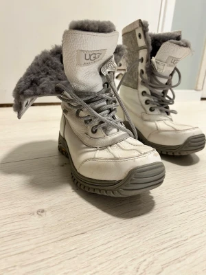 Vita vinterboots från UGG med ullfoder.  - Säljer ett par vita UGG boots med grått ullfoder och snörning. Skorna har robust sula, rund tå och detaljer i både skinn och textil. Går att använda med både nedvikta kanter eller som hög rak känga . Riktigt varma och sköna! Skorna är knappt använda! 