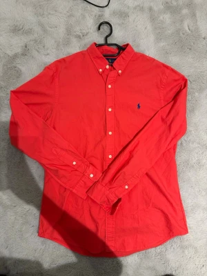 Röd skjorta från Ralph Lauren - Klassisk röd skjorta från Ralph Lauren i slim fit med blå broderad logga på bröstet. Skjortan har långa ärmar, knappar framtill och button-down krage. Tillverkad i bomull för en skön känsla och snygg passform.