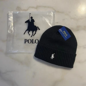 Svart stickad mössa från Polo Ralph Lauren - Svart stickad mössa från Polo Ralph Lauren med klassisk broderad logga i vitt framtill. Mössan har en bred ribbad kant och är tillverkad i mjukt material som håller dig varm. Perfekt accessoar för kyliga dagar och snygg till streetwear.