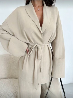 Beige set med knytband - Säljer denna beiga set med knytband i midjan och bred krage. Koftan har långärmade, lösa ärmar och är tillverkad i ett mjukt, strukturerat material som ger en chill och stilren vibe. Samma med byxorna. Den är helt ny, säljer den för den vart lite för stor på mig.