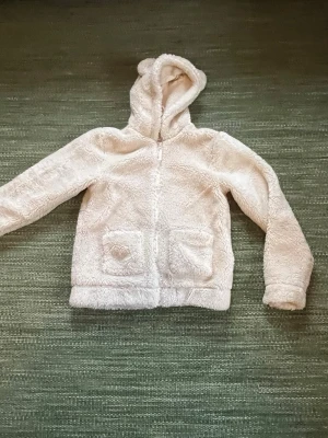 Beige teddyjacka med huva från H&M - Mysig beige teddyjacka från H&M med huva och dragkedja framtill. Jackan har två stora fickor fram och är tillverkad i fluffigt teddy-material som känns riktigt skönt. Perfekt för kyliga dagar och ger en avslappnad vibe.
