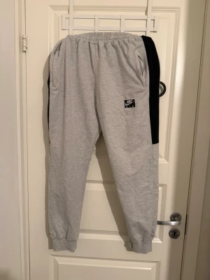 Nike Air grå mjukisbyxor - Snygga grå mjukisbyxor från Nike Air med svart sidopanel och broderad logga på benet. Byxorna har elastisk midja, muddar vid benslut och två fickor. Perfekta för chill eller träning, riktigt sköna och stilrena.