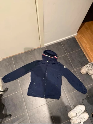 Marinblå vindjacka från Moncler - Säljer en marinblå vindjacka från Moncler med huva och dragkedja framtill. Jackan har Moncler-logga på ärmen och vita detaljer på fickorna. Materialet är lätt och syntetiskt, perfekt för blåsiga dagar. Snygg passform och klassisk design.