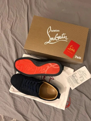 Mörkblåa Louboutins - Säljer ett par svarta sneakers från Christian Louboutin i mocka med den ikoniska röda sulan. Skorna har rund tå, svarta skosnören och beige läderfoder. Kommer med originalkartong, dustbag och kvitto. Perfekta för dig som vill sticka ut med exklusiv stil.