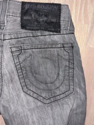 True Religion grå jeans bootcut - Säljer ett par gråa True Religion jeans med ikoniska kontrastsömmar och stora bakfickor. Jeansen har bootcut passform och klassisk femficksdesign. Tillverkade i USA och har en snygg tvättad look med svarta detaljer och knappgylf.