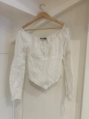 Vit blus med spets och knyt - Romantisk vit blus med lång ärm, knyt i halsringningen och spetsdetaljer vid ärmslut och nederkant. Tyget har diskreta prickar i strukturen och blusen har en lätt v-ringad offshoulder-modell. Perfekt för en somrig look.