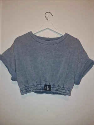 Blå croppad topp från Calvin Klein - Trendig croppad sweatshirt från Calvin Klein. Den har en avslappnad passform med rullade ärmar och en elastisk nederkant med en liten CK-logga. Snygg till träning, dans eller avslappnad stil.