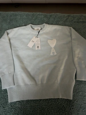 Ljusblå stickad tröja från Ami - Säljer en ljusblå stickad tröja från Ami Alexandre Mattiussi med ikoniskt hjärta och A på bröstet. Tröjan har rund hals, ribbade muddar och är långärmad. Tillverkad i mjuk bomull för en clean och chill vibe.