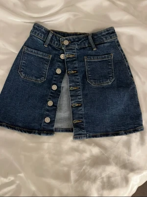 Mörkblå jeanskjol med knappar - Snygg mörkblå jeanskjol i kort modell med raka linjer och två stora fickor framtill. Kjolen har en rad med silverfärgade knappar längs hela framsidan och bälteshällor i midjan. Perfekt för dig som gillar en klassisk denimlook.