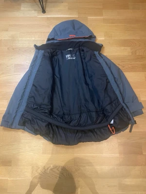 Mörkgrå Helly Hansen vinterjacka - Säljer en mörkgrå vinterjacka från Helly Hansen med avtagbar huva och orange HH-logga. Jackan har dragkedja framtill, två sidofickor med dragkedja och justerbara muddar vid ärmsluten. Perfekt för kalla dagar och riktigt snygg streetstil.